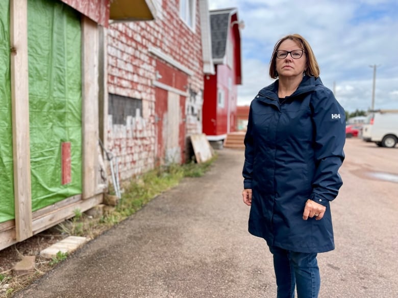 A woman in a long blue raincoat stands on a fishing wharf with damaged buildings in the background.