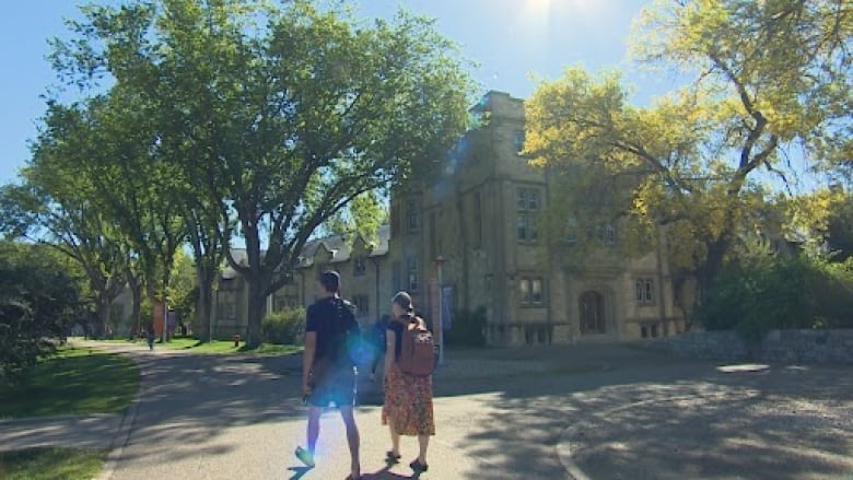 Two students walk under trees at a university campus with a sundog descending from the blue skies