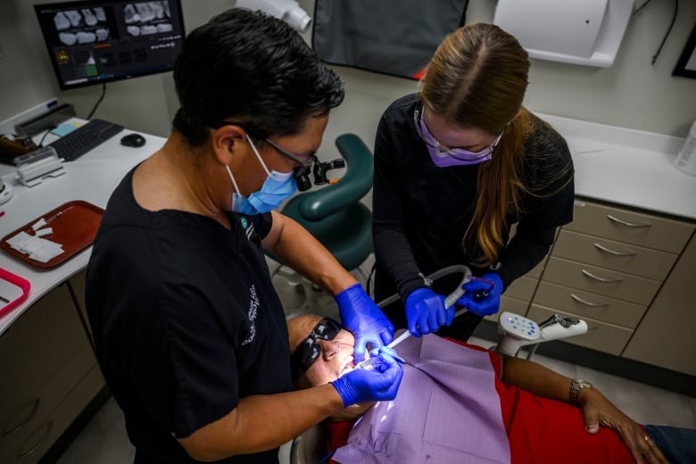 Two dental care professionals stand above a patient. Dental implements are in the patients mouth.