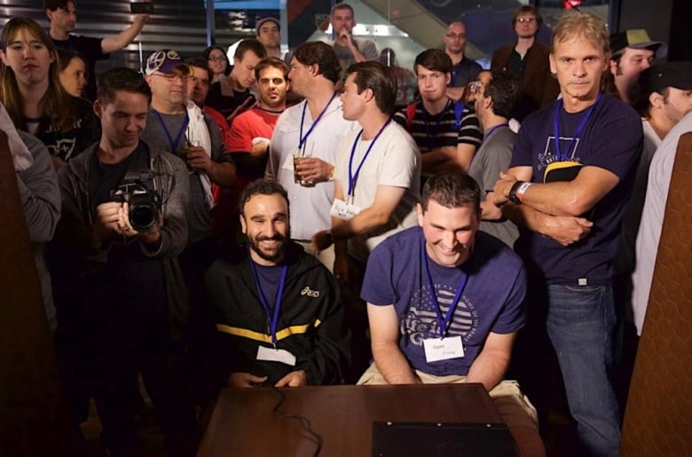 A crowd of onlookers, including one holding a camera, watches as two smiling men play a hockey video game at a tournament.