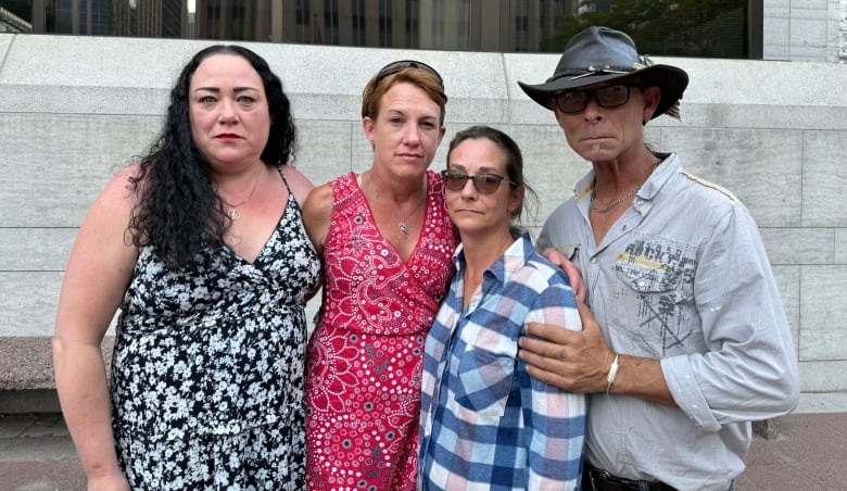 Four people with neutral expressions stand together in front of a stone building.