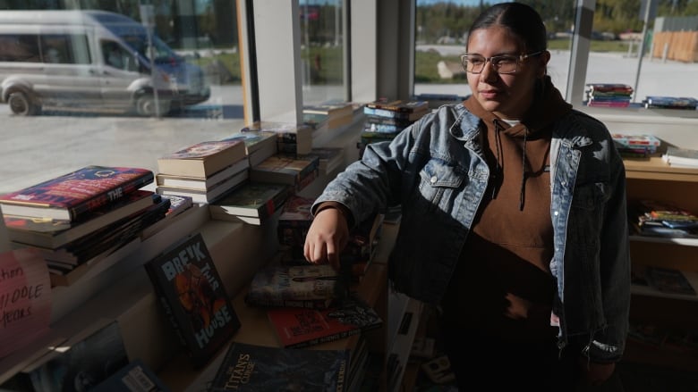 A student stands by a book shelf in a school library with windows in the background.