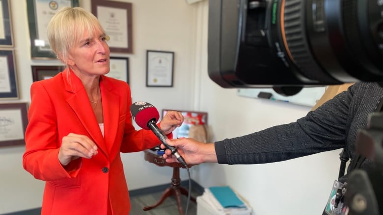 A woman with blonde hair wearing a red suit speaks into a microphone and camera in an office.