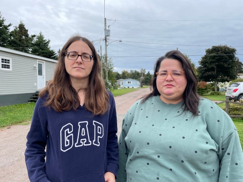 Two women stand outside for a photo with straight faces. There are mobile homes behind them. They are in a neighborhood. Both have brown hair and blue sweatshirts. 