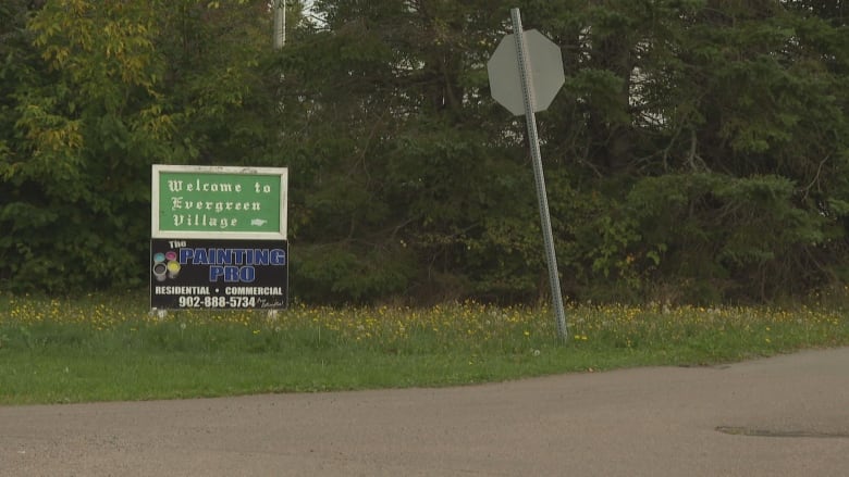 A green sign that says Evergreen Village. It is on the grass near a quiet street. There are trees behind it. 