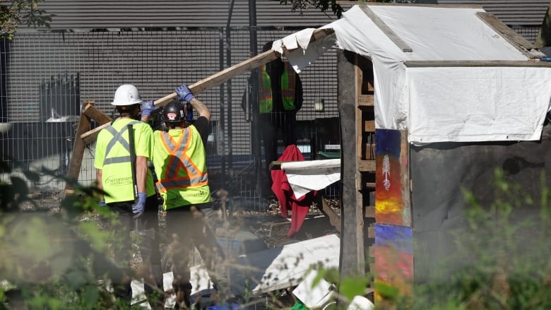 Two men in high-visibility vests use sledgehammers and a pry bar to tear down a shed-like shelter.