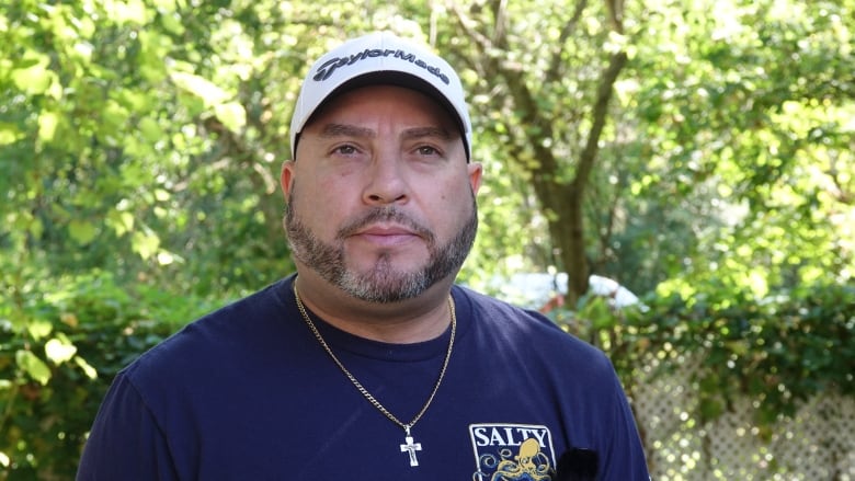 A man with a chin-strap beard wearing a ball cap stares at the camera. The background is leafy, green trees and sunshine.