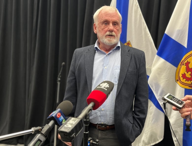 A man with white hair and beard stands in front of microphones.