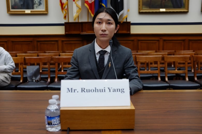 An oriental man with long black hair, tied in a pony tail, dressed in a grey three-piece suit, sits at a microphone with empty chairs behind him,