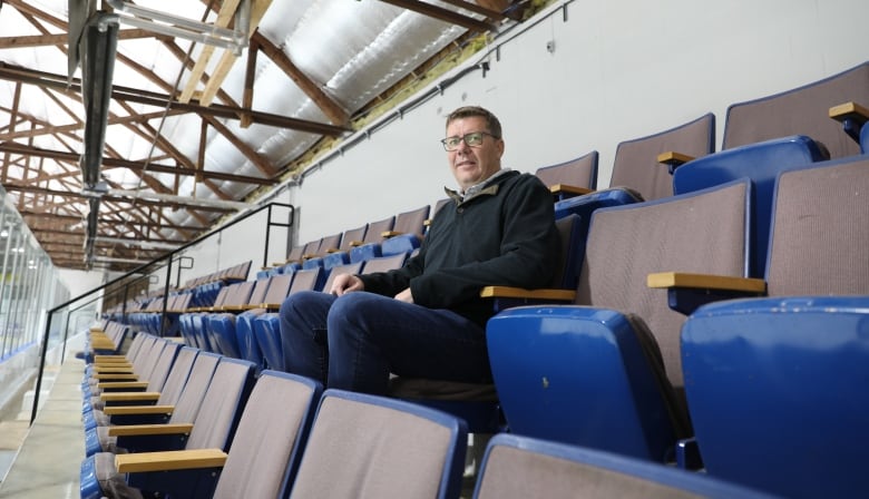 A man in a collared sweater sits in a hockey rink seat