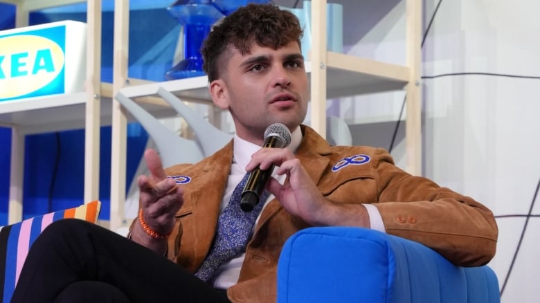 A man in a suit and tie sits in a chair and speaks into a microphone.