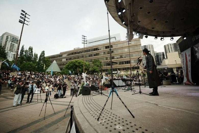 A large crowd gathers in front of an outdoor stage. A man in a black trenchcoat and black boots speaks at a microphone.