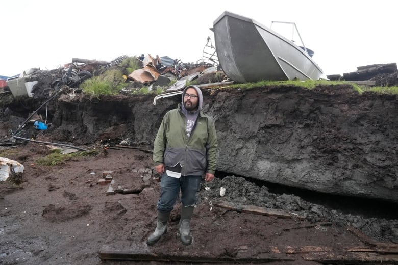 A man stands by a steep muddy bank piled with debris.