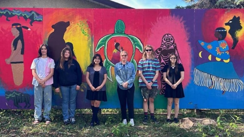 A group of students leans against a multi-coloured mural that is hung on a fence near a path. 