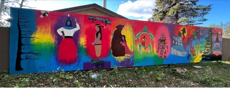 A colourful mural with multiple panels leans against a fence near a walking path outside. 