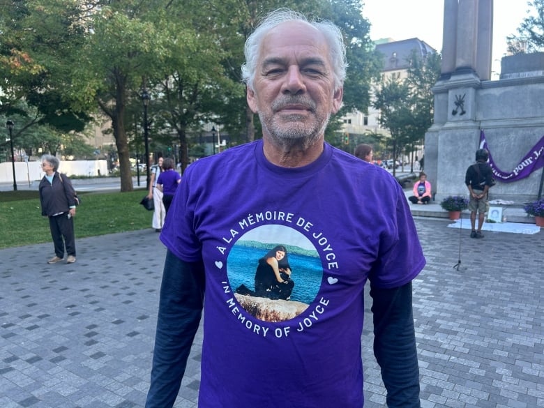 A man with grey hair. He's wearing a purple shirt with the picture of a woman sitting on a rock by a lake. Around the picture are the words: 