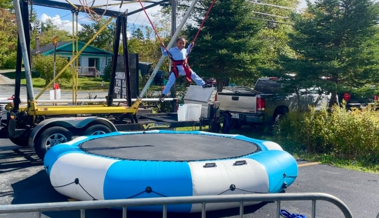 A young girl is jumping on a trampoline outside. She is wearing a harness. 