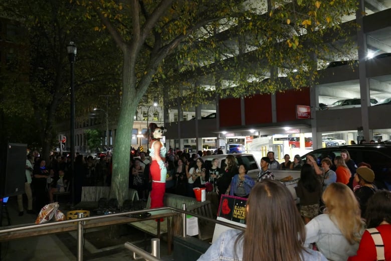 A person stands on a bench dancing in front of a crowd outside at night. 