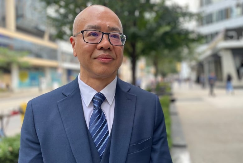 A man with glasses wearing a suit stands on the sidewalk.