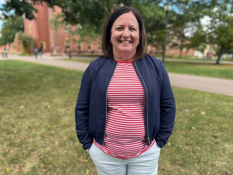 A woman with a blue coat and red stripped top smiles for the camera while on a university campus with trees and brick buildings behind her. 