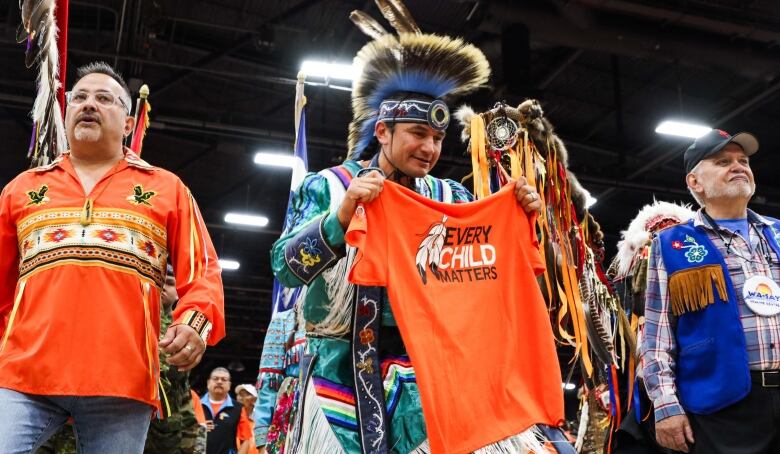 A man wearing powwow regalia hold an orange shirt that says 'Every Child Matters'