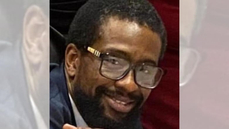 A black man with a beard and glasses smiles at the camera. He's dressed in blue and only his head is visible