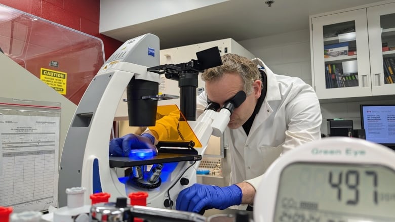 Man in white lab coat looking through microscope-type device.