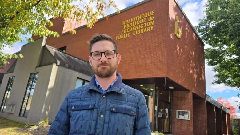 An unsmiling man with glasses standing in front of a library.