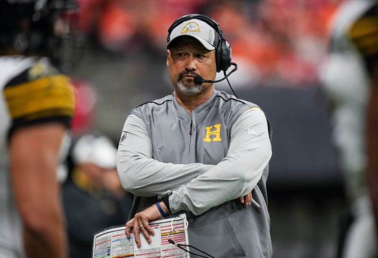 A football couch stands on the sidelines wearing a headset while watching his team players during a game. 