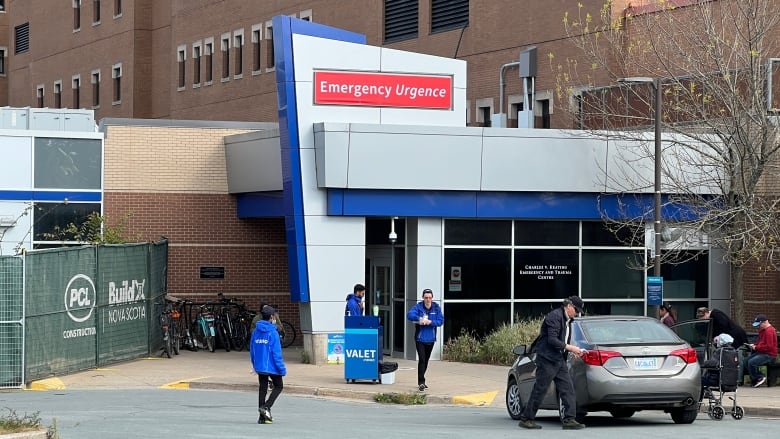 Three workers in blue, stand near a Valet sign, in front of an emergency department at a Halifax hospital. On the right, a grey car can be seen, with the driver seen exiting the vehicle, and a passenger in a wheelchair on the right. 