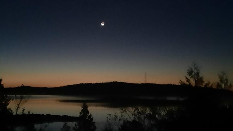 The moon glows over a brightening horizon and river.