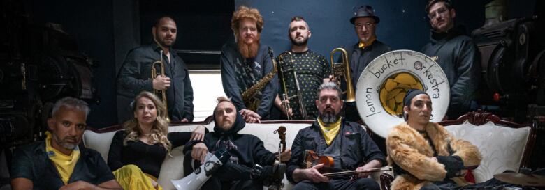 Ten members of an orchestra called  Lemon Bucket Orkestra pose for a photo holding instruments from trumpets to violins. Five members sitting on a couch, five standing behind.