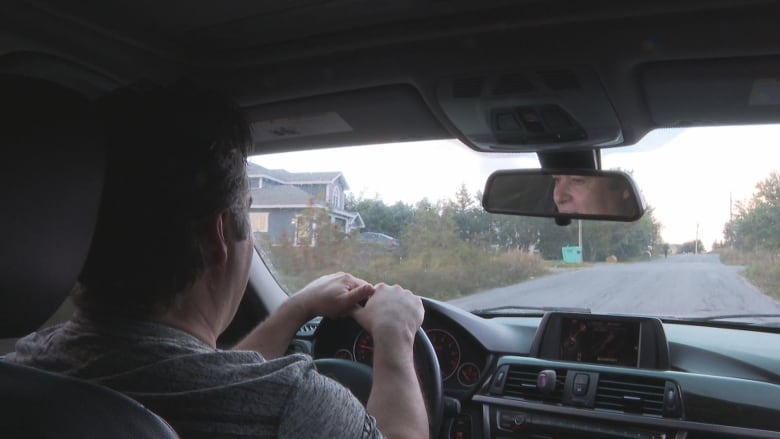 A man is seen driving inside of a car. His face is visible in the rearview mirror. Both of his hands are on the steering wheel.