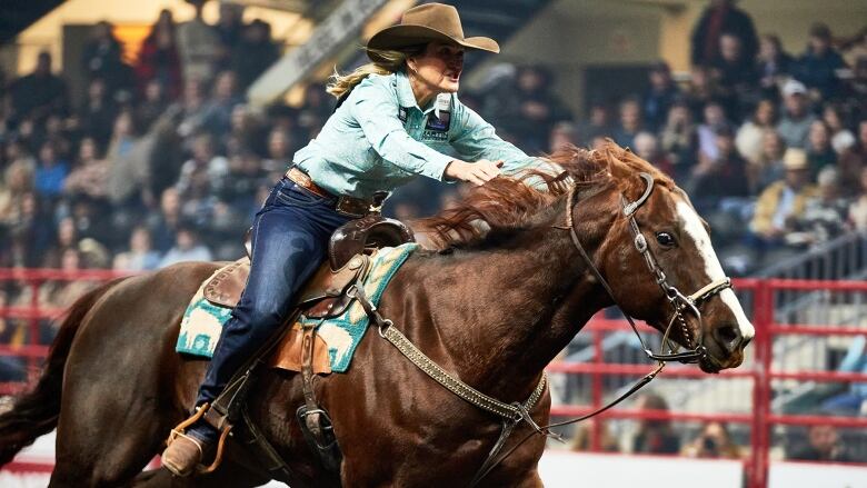 A woman in a cowboy hat rides a sorrel-colored horse in an arena.