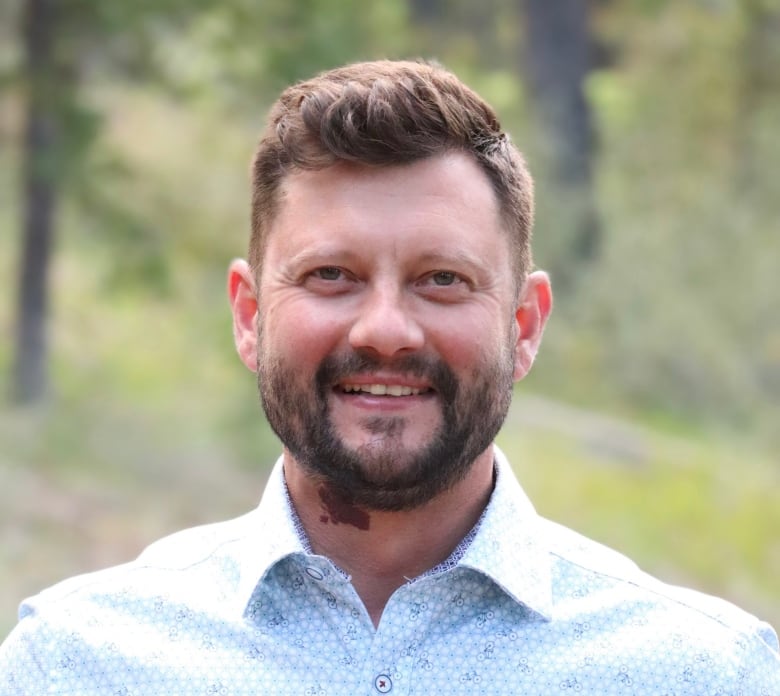 A man smiles in front of a wooded background.