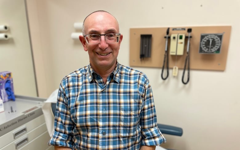 Man with glasses and blue plaid shirt stands in a hospital clinic room. 