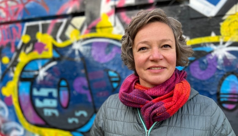 A woman in a puffy jacket stands against a graffiti'd wall, looking cheerful. 