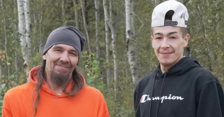 A man wearing an orange hoodie with a 'W' on it and a toque, stands screen left to another man wearing a black hoodie and a backwards hat as they pose for the camera. 