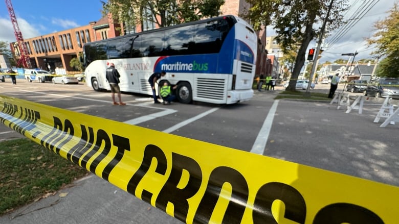 Yellow police tape in the foreground with a motorcoach bus in the background and two people crouching down by one of the tires.