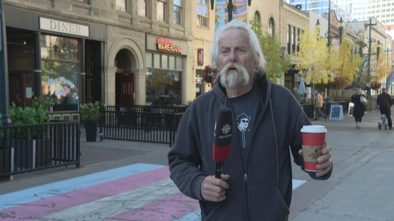 a man with a beard holding a coffee cup speaks into a microphone. he stands on a busy pedestrian street.