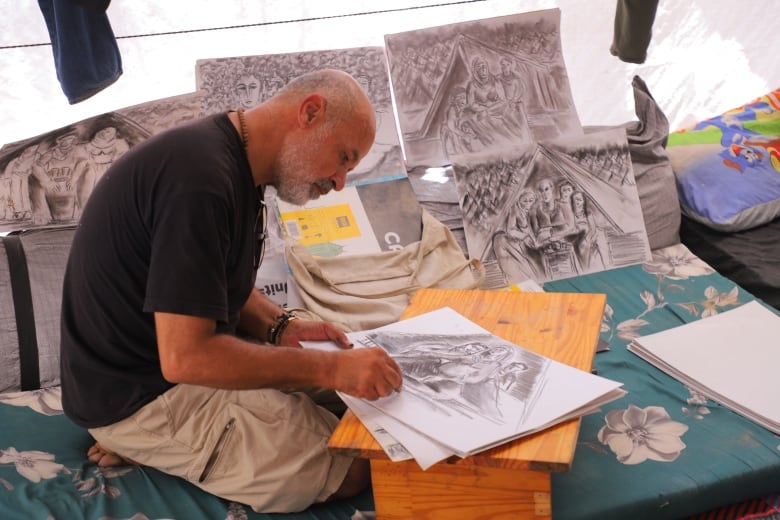 Man sitting on the floor inside a makeshift tent, drawing.