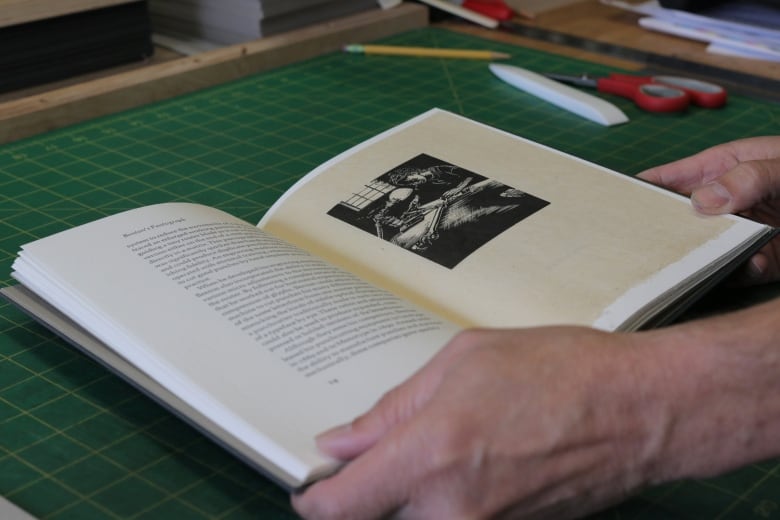 A book faces is held open by a pair of hands on a table. Scissors and a pencil are nearby.