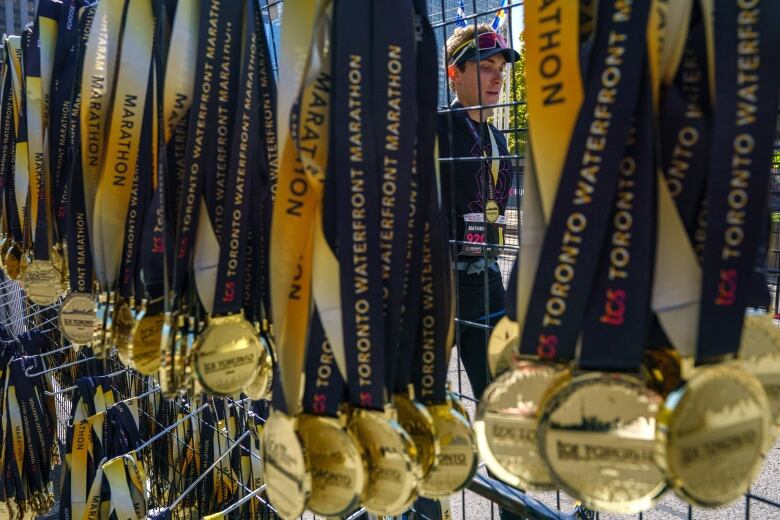 Dozens of gold medals hand from a chain fence. A man in a racing bib with a medal around his neck is visible partly through the fence