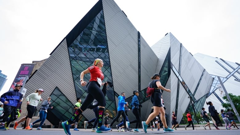 On an overcast fall day, dozens of runners with racing bibs run on a city street passing the glass, Royal Ontario Museum