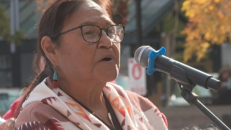 A woman with brown hair wearing glasses speaks into a microphone.