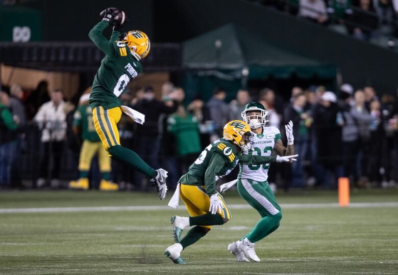 A football player in a green-and-yellow uniform is in mid-air, intercepting a pass. Two other players - a teammate and an opponent - are further down the field, watching.