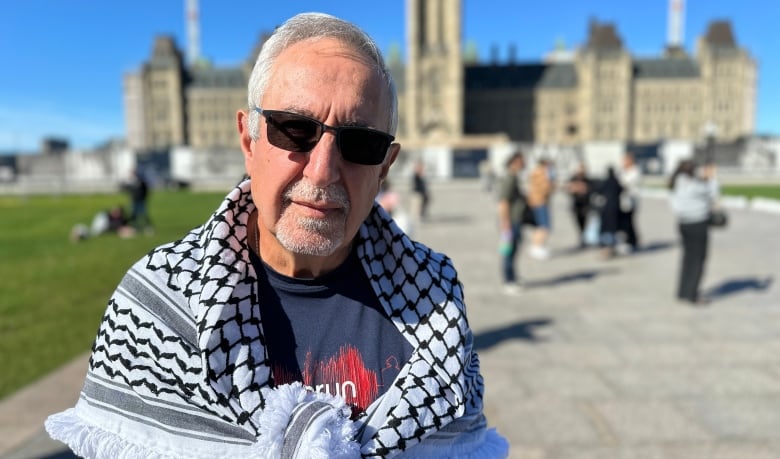 A man with white hair and sunglasses stands in front of Canada's parliament with a Keffiyeh draped around his shoulders.