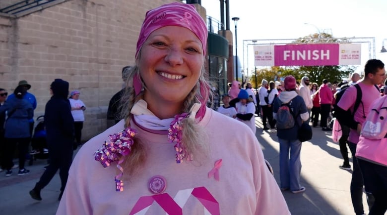 A woman wearing a pink bandana and shirt smiles to the camera as people gather in front of a finish line behind her.