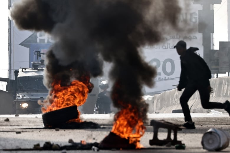 Large black clouds rise to the sky in an urban setting from orange fires erupting on the ground from burnt tires. To the right, a person in shadow is shown.