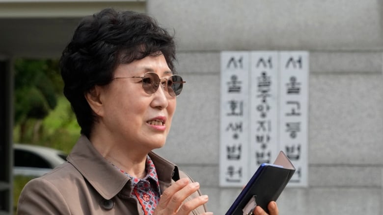 An older, dark haired Asian woman wearing glasses appears to speak into a phone while standing outside.